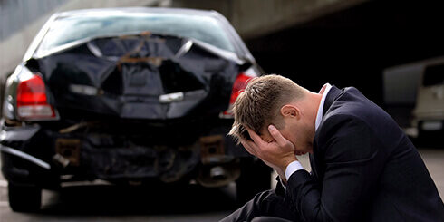 Photo of a man sitting on a curb with his head in his hands beside a car that has been rear ended